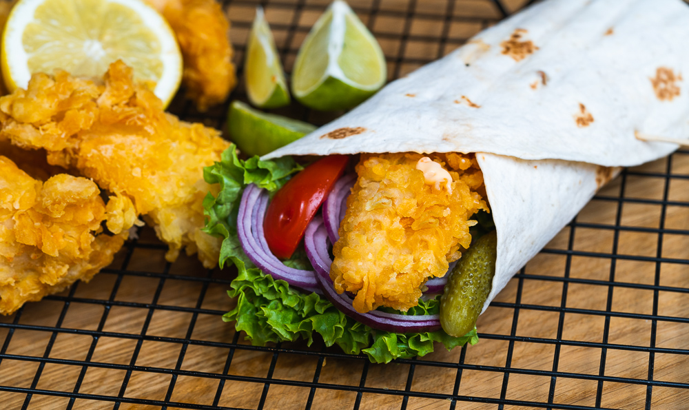 un wrap de pollo y verduras con trozos de pollo crujiente a su lado y unos cuartos de limón