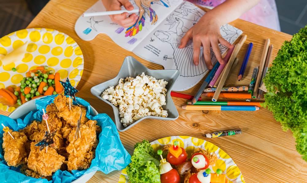 una niña está dibujando sentada en la mesa llena de pollo frito y verduras